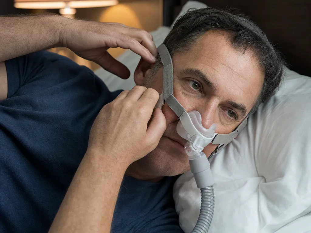 Hände justieren im Liegen das Kopfband einer CPAP-Nasenmaske nach, um ein Leck am Maskenrand zu schließen
